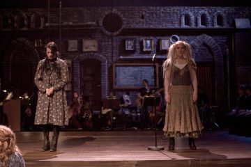 Kaksi näyttelijää seisoo hämärästi valaistulla näyttämöllä. Toinen, tummahiuksinen, katsoo alaspäin kädet ristissä, kun taas toinen, vaaleahiuksinen, seisoo mikrofonin vieressä ja katsoo eteenpäin. Taustalla tiiliseinää vasten näkyy orkesteri.