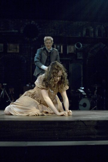 Vaaleanruskeaan mekkoon pukeutunut nainen polvistuu näyttämön lattialla ahdistuneen näköisenä, ja tummapukuinen mies seisoo hänen takanaan huolestunut ilme kasvoillaan. Taustalla on hämärä valaistus, jossa näkyy soittimia.