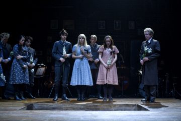 Lavalla seisoo joukko juhlallisia, vintage-tyylisesti pukeutuneita ihmisiä, joilla on kädessään valkoisia kukkia ja jotka ovat kerääntyneet suorakulmaisen avoimen haudan tai kuopan ympärille hämärässä valaistussa ympäristössä, mikä viittaa hautajaisiin tai muistotilaisuuteen.