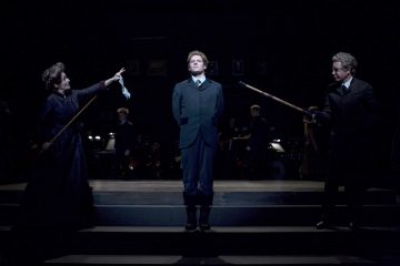 Kolme näyttelijää seisoo hämärästi valaistulla näyttämöllä; keskimmäinen hahmo seisoo liikkumatta kädet selän takana, kun taas kaksi näyttelijää molemmin puolin osoittaa häntä pitkillä keppeillä, mikä luo jännittyneen, dramaattisen kohtauksen. Taustalla näkyy heikosti muusikoita.