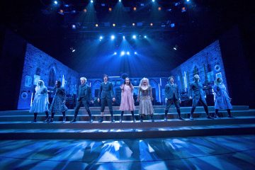 Ryhmä pukuihin pukeutuneita näyttelijöitä seisoo rivissä lavalla, jossa on sininen valaistus, ja esiintyy tiiliseinäistä huonetta muistuttavan lavastuksen edessä.