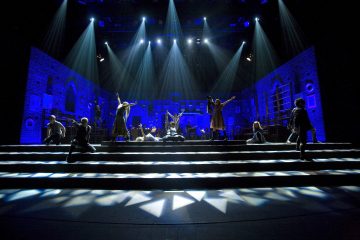 Ryhmä esiintyjiä teatterilavalla, jossa on sininen valaistus, dramaattiset valonheittimet ja kaupunkimaisemaa muistuttava lavastus. Osa esiintyjistä seisoo kädet kohotettuna, osa istuu tai polvistuu portailla.