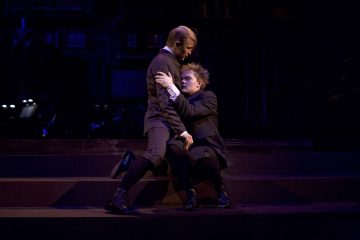 Lavalla esiintyy kaksi näyttelijää vanhanaikaisissa, tummissa juhlavaatteissa. Toinen polvistuu ja tarrautuu toiseen, joka seisoo ja katsoo häntä alaspäin. Valaistus on dramaattinen, mikä korostaa heidän tunnepitoisia ilmeitään.