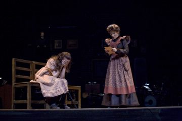 Lavalla kaksi naista aikakauden puvuissa; toinen istuu penkillä kasvot käsissään ja vaikuttaa ahdistuneelta, kun taas toinen seisoo ja lukee paperia huolestuneen näköisenä. Tausta on tumma ja teatraalinen.