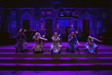 Seitsemän nuorta naista laulaa lavalla istuen ja polvistuen, intensiiviset ilmeet kasvoillaan, kun taas neljä tummiin asuihin pukeutunutta nuorta miestä seisoo heidän takanaan. Dramaattinen lavastus on valaistu violetilla, ja siinä on tiilikaaria ja kehystettyjä kuvia.