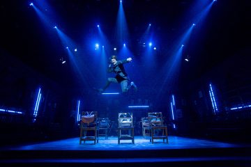 Tummiin vaatteisiin pukeutunut esiintyjä hyppää keskellä ilmaa näyttämöllä olevan tuolirivin yläpuolella, jota valaisee dramaattinen sininen valaistus teatteriympäristössä.