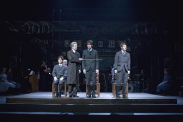 Näyttämökohtaus, jossa on neljä näyttelijää aikakauden puvuissa; yksi vanhempi mies seisoo keppi kädessään ja puhuttelee kahta nuorta miestä, kun toinen istuu lähellä. Taustalla näkyy muusikoita ja muita näyttelijöitä. Lavastus muistuttaa tiiliseinäistä luokkahuonetta.