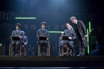 Neljä vanhanaikaisiin koulupukuihin pukeutunutta nuorta miestä istuu näyttämöllä pulpettien ääressä. Heidän vieressään seisoo vanhempi mies pitkässä takissa, jolla on keppi kädessään ja joka puhuu yhdelle oppilaalle. Lava on hämärästi valaistu sinisellä hehkulla ja neonvihreillä valoilla.