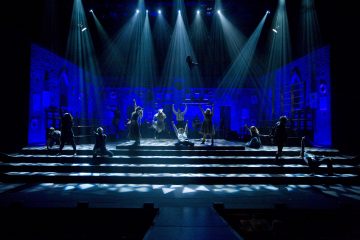 Esiintyjät ovat dramaattisesti poseeranneet lavalla, jossa on sininen valaistus ja valonheittimet, ja taustalla on tiiliseinä ja goottilaisia ikkunoita, jotka luovat teatraalisen ja dynaamisen ilmapiirin.