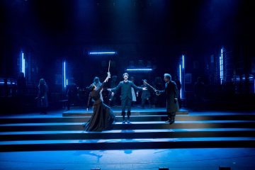 Dramaattisessa, sinisellä valaistussa näyttämökohtauksessa näkyy tummiin vaatteisiin pukeutunut mies, joka seisoo portailla kädet ojennettuina, kun kaksi ihmistä osoittaa häntä sauvoilla. Taustalla seisoo useita näyttelijöitä hehkuvien pystysuorien valaisimien keskellä.