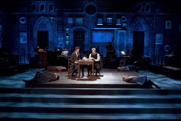 Ryhmä miehiä istuu pöydän ääressä lavalla.