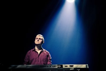 Punapaitainen henkilö seisoo näppäimistön takana pimeällä lavalla, jota valaisee ylhäältä alaspäin loistava kirkas valonheitin.