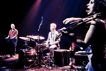 Lavalla esiintyy kolme muusikkoa dramaattisessa valaistuksessa: yksi soittaa koskettimia, toinen rumpuja ja kolmas etualalla soittaa energisesti viulua. Taustalla on pimeää ja värillisiä näyttämövaloja.