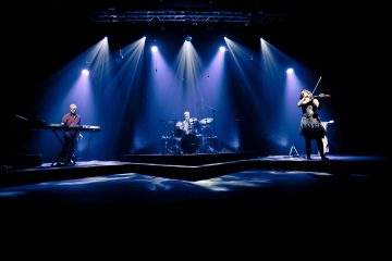 Bändi esiintyy tummalla lavalla, jossa on sinisiä valonheittimiä; kosketinsoittaja seisoo vasemmalla, rumpali keskellä takana ja viulisti oikealla, ja kaikki soittavat instrumenttejaan.