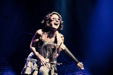 Silmälasipäinen nainen hymyilee leveästi soittaessaan sähköviulua lavalla, jota valaisee dramaattinen sininen ja violetti valaistus.