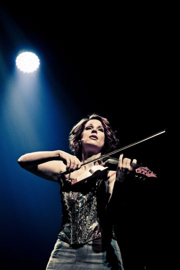 Kimaltavaan korsettiin pukeutunut nainen soittaa sähköviulua näyttämöllä dramaattisen valokeilan alla tummalla taustalla.