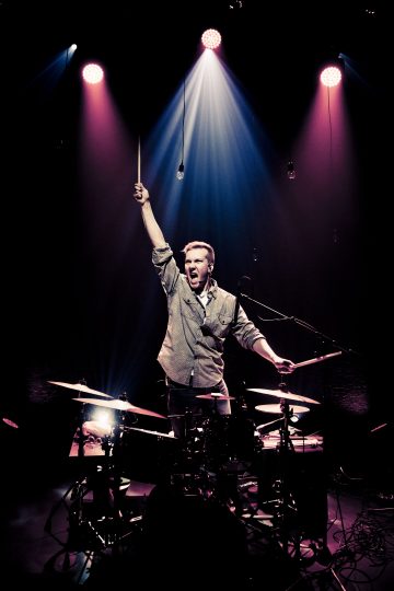 Rumpali seisoo rumpukaluston takana pimeällä lavalla, nostaa yhden rumpukapulan korkealle, suu auki kuin huutaisi, ja alaspäin loistavat dramaattiset punaiset ja siniset valonheittimet.