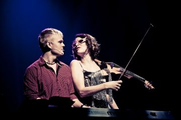 Punaruutuinen mies soittaa koskettimia punaisessa ruutupaidassa viulua soittavan, kimaltelevaan toppiin ja aurinkolaseihin pukeutuneen naisen vieressä, ja molemmat ovat lavavalojen valaisemia tummaa taustaa vasten.