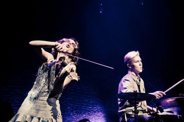 Nainen soittaa intohimoisesti sähköviulua, kun hänen vieressään oleva mies soittaa rumpuja lavalla, ja molemmat ovat dramaattisen näyttämövalaistuksen valaisemia tummaa taustaa vasten.