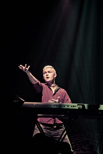 Lyhyet vaaleat hiukset omaava henkilö soittaa lavalla näppäimistöä, hänellä on punainen paita yllään ja hän elehtii dramaattisesti vasemmalla kädellään värikkäässä valaistuksessa.