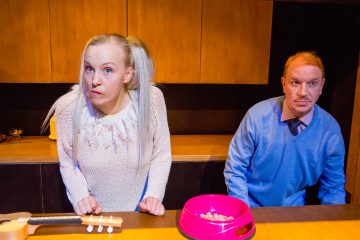 Nainen, jolla on vaaleat letit ja yllättynyt ilme, nojaa tiskipöydälle siniseen puseroon pukeutuneen, vakavalta näyttävän miehen viereen. Heidän edessään tiskillä on vaaleanpunainen lemmikkikulho ja pieni ukulele.