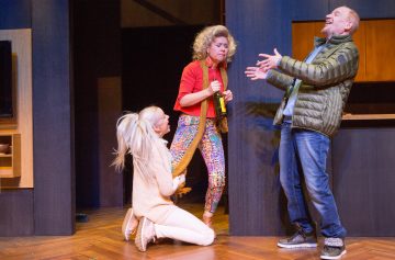 Lavalla esiintyy kolme näyttelijää: yksi nainen polvistuu ja tarttuu toisen naisen jalkaan; toinen nainen seisoo ja pitää pulloa kädessään ja näyttää vakavalta; vihreätakkinen mies reagoi yllättyneenä, kädet levällään.