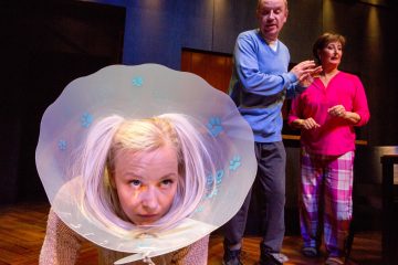 Lattialla kyyristelee vaaleahiuksinen nainen, jolla on vaaleat hiukset letitettyinä, muovinen lemmikkikartio päässään. Hänen takanaan siniseen villapaitaan pukeutunut vanhempi mies ja vaaleanpunaiseen pyjamaan pukeutunut nainen katsovat hämmästyneinä.