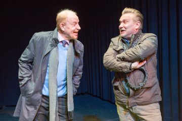 Kaksi miestä seisoo lavalla tummien verhojen edessä ja keskustelee vilkkaasti. Toisella on takki ja huivi päällään ja hän hymyilee lämpimästi, kun taas toinen seisoo kädet ristissä ja vaikuttaa huvittuneelta. Molemmilla on vaaleat hiukset ja ilmeikkäät kasvot.