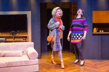 Kaksi naista seisoo ja nauraa kirkkaasti valaistussa olohuoneessa. Toisella on päällään kuviollinen takki, värikkäät housut ja saappaat, kun taas toisella on raidallinen mekko, jossa on pilkkuja. Taustalla näkyy sohva ja hyllyjä.