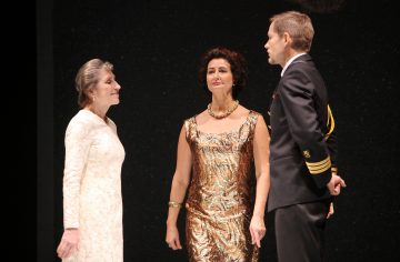 Lavalla seisoo kolme ihmistä: vanhempi nainen pitkässä valkoisessa mekossa, nainen kultakuvioisessa mekossa ja kaulakorussa sekä mies mustassa sotilasunivormussa, jotka kaikki näyttävät keskustelevan.
