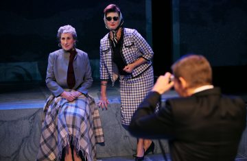 Kaksi vintage-asuihin pukeutunutta naista poseeraa valokuvaa varten; toinen istuu ruudullinen huopa sylissään, toinen seisoo vieressä aurinkolasit ja huivi päällään. Pukuun pukeutunut henkilö ottaa kuvan teatteritilassa.