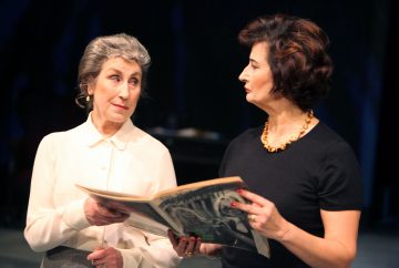 Kaksi naista seisoo tiiviisti vierekkäin; toisella valkoisessa puserossa on avoin sanomalehti kädessään, kun taas toinen mustassa topissa ja kultaisessa kaulakorussa katsoo häntä mietteliäänä. Tausta on tumma ja epätarkka.