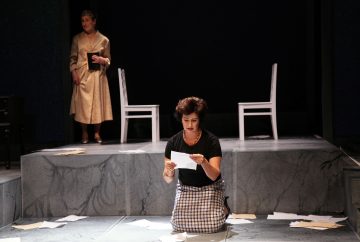 Nainen polvistuu lavalla ja lukee paperia, jota ympäröivät hajallaan olevat arkit, kun taas toinen nainen seisoo taustalla pitelemässä kirjaa. Heidän takanaan on kaksi valkoista tuolia. Kohtaus on dramaattinen ja teatraalinen.