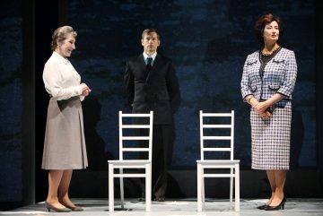 Kolme näyttelijää näyttämöllä: kaksi vintage-tyyliseen asuun pukeutunutta naista seisoo valkoisten tuolien vieressä vastakkain, ja tummapukuinen mies seisoo heidän välissään ja takanaan tummaa taustaa vasten.