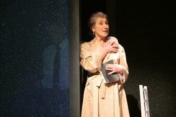 Vanhempi nainen seisoo beigeen kaapuun pukeutuneena, kirja kädessään ja rintaansa puristaen, mietteliään näköisenä. Hän seisoo lähellä tummaa seinää, jossa on heikko heijastus, ja hänen vieressään näkyy valkoinen tuoli.