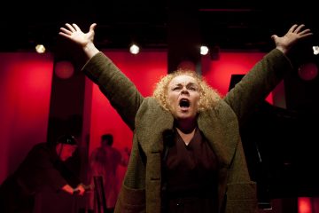 Nainen, jolla on kiharat vaaleat hiukset, seisoo lavalla kädet kohotettuna ja suu auki kuin huutaen. Hänellä on ruskea takki. Taustalla on punainen dramaattinen valaistus, ja taustalla on toinen henkilö pianon äärellä.