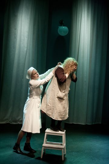Valkoiseen univormuun pukeutunut hoitaja avustaa askelmatuolilla seisovaa kiharatukkaista naista, joka on käärinyt vaatteen hartioilleen, ja taustalla on pehmeästi valaistu näyttämö, jossa on korkeat verhot.