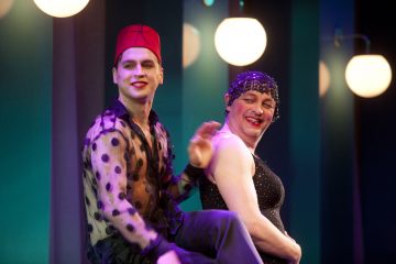 Kaksi näyttäviin pukuihin ja meikkiin pukeutunutta henkilöä esiintyy lavalla lämpimien valojen alla. Toisella on punainen hattu ja läpikuultava pilkullinen toppi, toisella on kimalteleva musta mekko ja päähine. Molemmat hymyilevät ja poseeraavat leikkisästi.