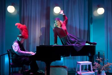 Punaiseen asuun ja höyhenpäähineeseen pukeutunut henkilö soittaa pianoa, kun taas toinen henkilö punaisessa takissa ja mustassa hameessa poseeraa dramaattisesti pianon päällä. Kohtaus on kirkkaasti valaistu sinisellä ja vaaleanpunaisella valaistuksella.