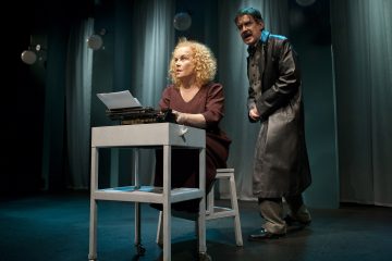 Kihara vaaleahiuksinen nainen istuu kirjoituspöydän ääressä ja kirjoittaa kirjoituskoneella, kun hänen takanaan seisoo tummiin pitkiin takkeihin pukeutunut mies, joka puhuu intensiivisesti. He ovat hämärästi valaistulla näyttämöllä, jossa on siniset verhot ja roikkuvat valot.