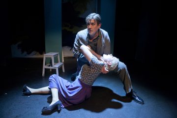 Mies polvistuu lavalla ja pitää sylissään dramaattisesti lyyhistynyttä naista. Naisella on violetti hame ja kuviollinen pusero; mies on pukeutunut harmaaseen. Heihin kohdistuu valonheitin, ja taustalla on pieni jakkara.