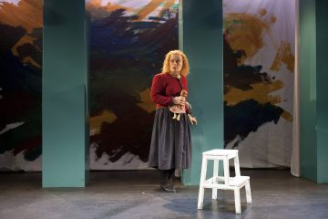 Punaiset kiharat hiukset omaava nainen seisoo lavalla nukke kädessään, yllään punainen pusero ja tumma hame. Hän seisoo tiilenpunaisen pylvään välissä, jonka taustalla on värikäs abstrakti tausta ja lähellä valkoinen askelmatikka.