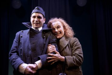 Sotilastyyliseen takkiin ja hattuun pukeutunut mies seisoo käsi kädessä hymyilevän takkiin pukeutuneen naisen kanssa, molemmat lämpimästi valaistuna pimeällä näyttämöllä. Nainen tarttuu hellästi miehen käteen, ja molemmat näyttävät tyytyväisiltä.