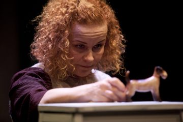 Punaiset kiharat hiukset omaava nainen katsoo keskittyneesti kädessään olevaa pientä koirahahmoa, joka on asetettu valkoiselle pinnalle tummaa taustaa vasten.