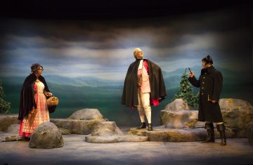 Kolme näyttelijää seisoo lavalla aikakauden puvuissa keinotekoisten kivien keskellä, ja taustalla on maalattu pilvinen taivas ja kukkuloita. Yhdellä näyttelijällä on viitta ja saappaat, kun taas muilla on viitta ja takki, jossa on hattu.