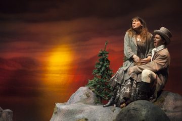 Lavalla esiintyy kaksi näyttelijää, jotka ovat pukeutuneet aikakauden pukuihin ja istuvat kivillä, joilla on pieni mänty. Taustalla näkyy dramaattinen auringonlasku, jossa on oranssin ja keltaisen sävyjä.