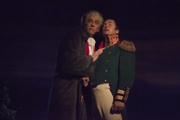 Kaksi teatteriasuihin pukeutunutta miestä seisoo lähekkäin hämärästi valaistulla näyttämöllä; toisella on harmaa turkistakki ja valkoinen peruukki, toisella vihreä sotilastakki, jossa on kultainen reunus ja olkaimet.