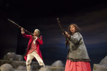 Kaksi näyttelijää näyttämöllä, toinen kirkkaanpunaisessa takissa ja valkoisissa housuissa ja toinen harmaassa takissa ja vaaleanpunaisessa hameessa, molemmat tähtäävät pitkillä kivääreillä kallioisen lavastuksen keskellä dramaattisessa valaistuksessa.