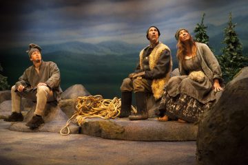 Kolme maalaismaisesti vanhanaikaisiin vaatteisiin pukeutunutta ihmistä istuu kivikkoisella maalla ja katsoo taivaalle. Heidän takanaan on mäntyjä ja heidän jalkojensa juurella on köysirulla, mikä viittaa ulkoilmaan, erämaiseen ympäristöön.