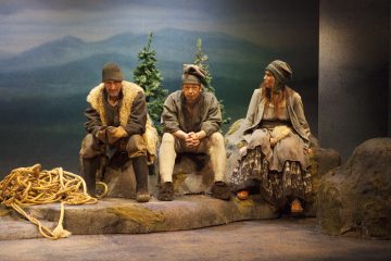 Kolme vanhanaikaisiin, riekaleisiin vaatteisiin pukeutunutta ihmistä istuu kivillä köyden vieressä, ja taustalla on maalatut vuoret ja puut, jotka viittaavat maaseutumaisuuteen tai historialliseen ympäristöön, mahdollisesti näyttämönäytelmästä.